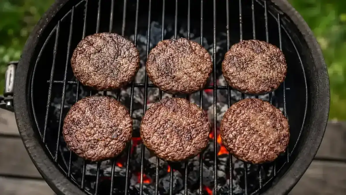 Six identical burger patties lined up on a grill grate cooking evenly over charcoal