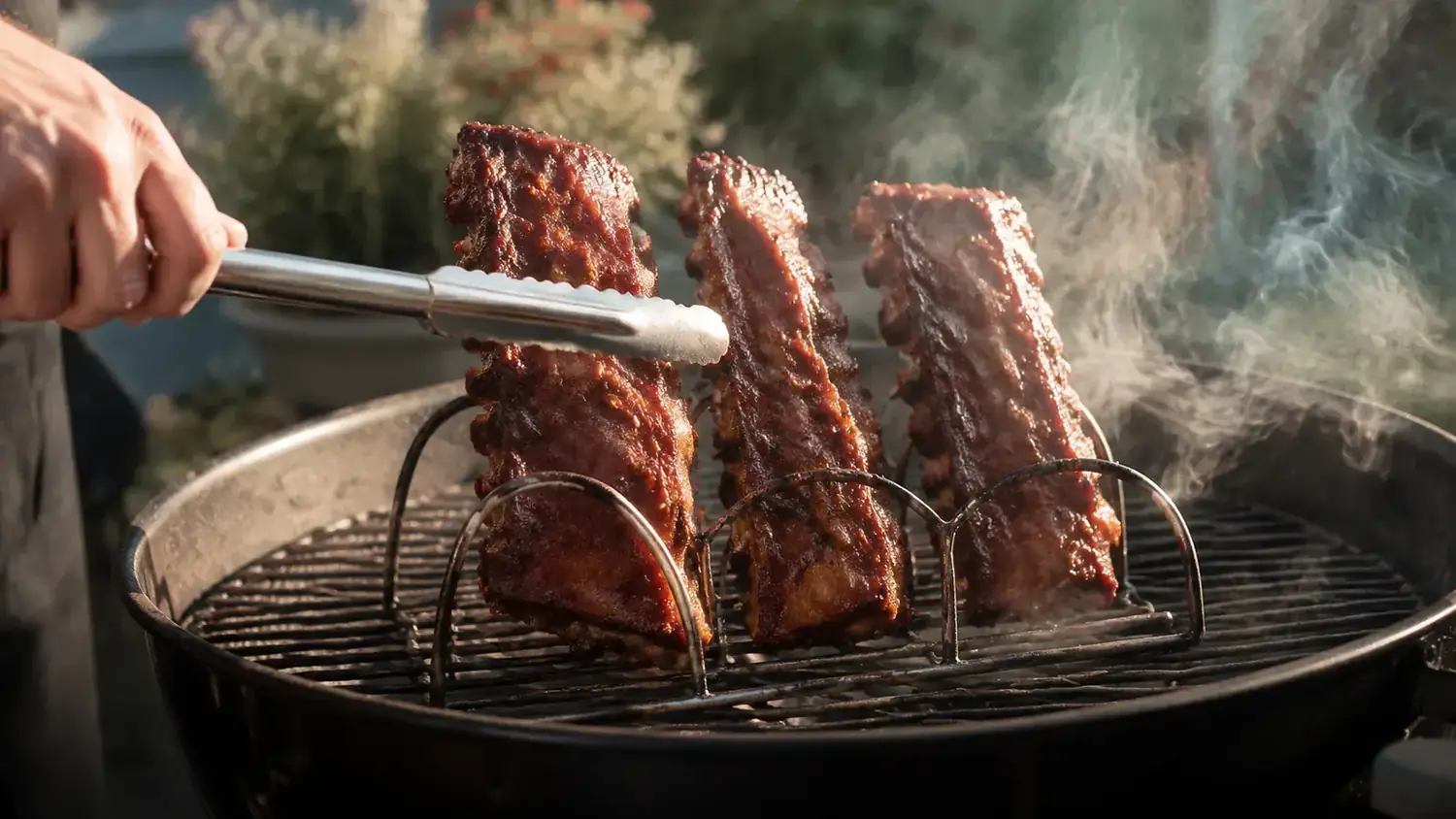 Three racks of baby back ribs standing upright in a stainless steel rib rack on a charcoal grill, smoke rising around them