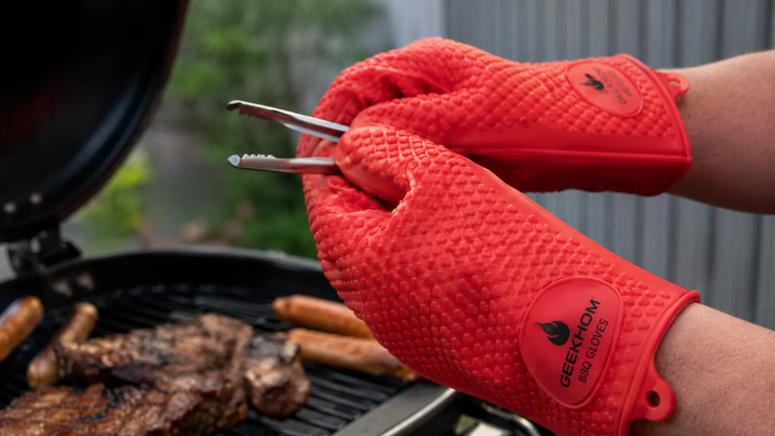 Hands wearing GEEKHOM silicone BBQ gloves attempting to pick up small tongs with a close-up showing limited finger dexterity