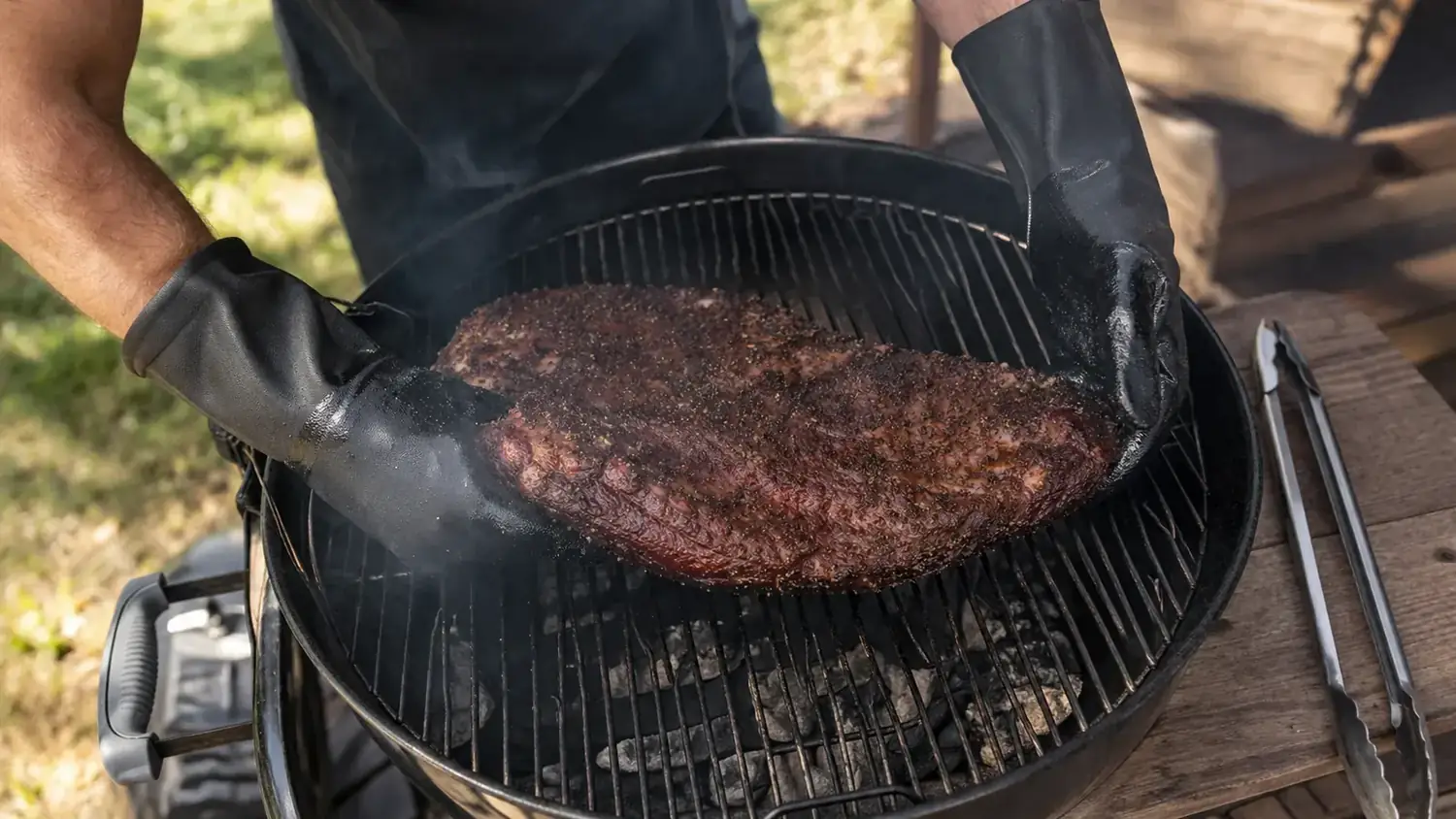 Backyard griller using BBQ gloves to flip a large brisket flat on a charcoal kettle grill