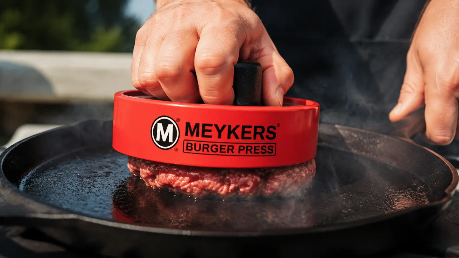 Meykers burger press being used to smash a beef ball on a hot cast-iron skillet