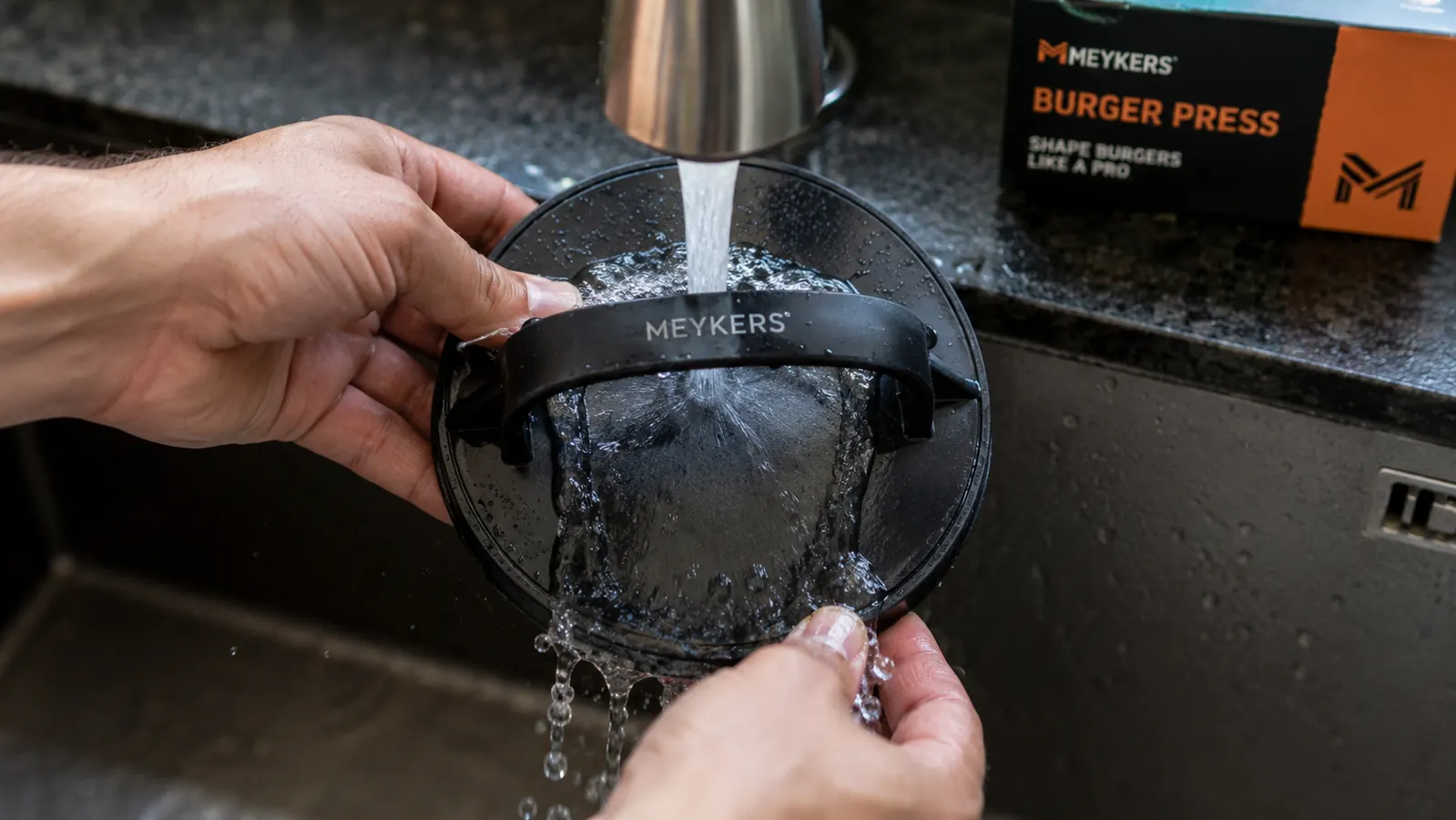 Meykers burger press being rinsed under a kitchen faucet, showing the non-stick surface with light staining