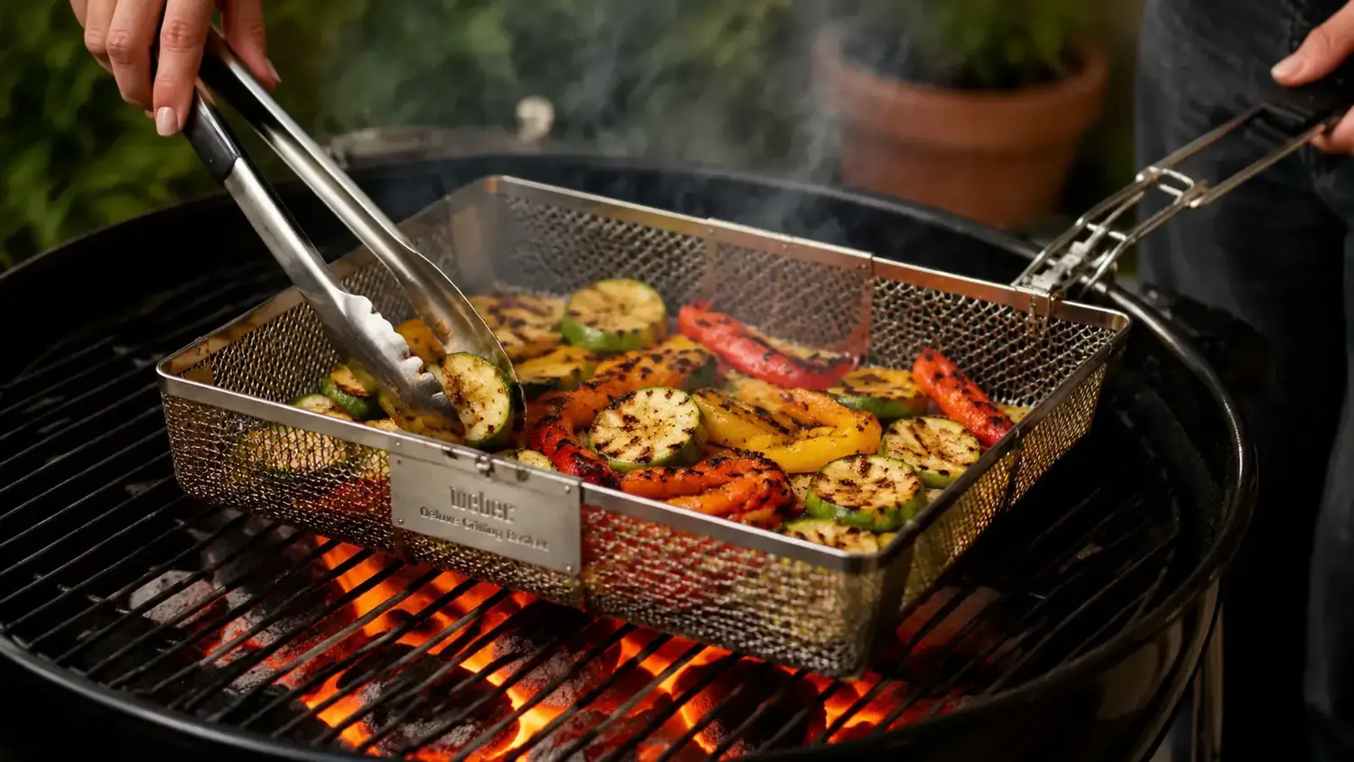 Hand using tongs to flip mixed vegetables inside the Weber Deluxe Grilling Basket over a charcoal fire